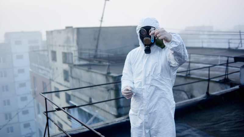 Focus on hands, holding glass flask with blue reagent. Scientific research on a subject environmental catastrophe, conducted by a scientist in white coverall and gas mask, working on roof of building